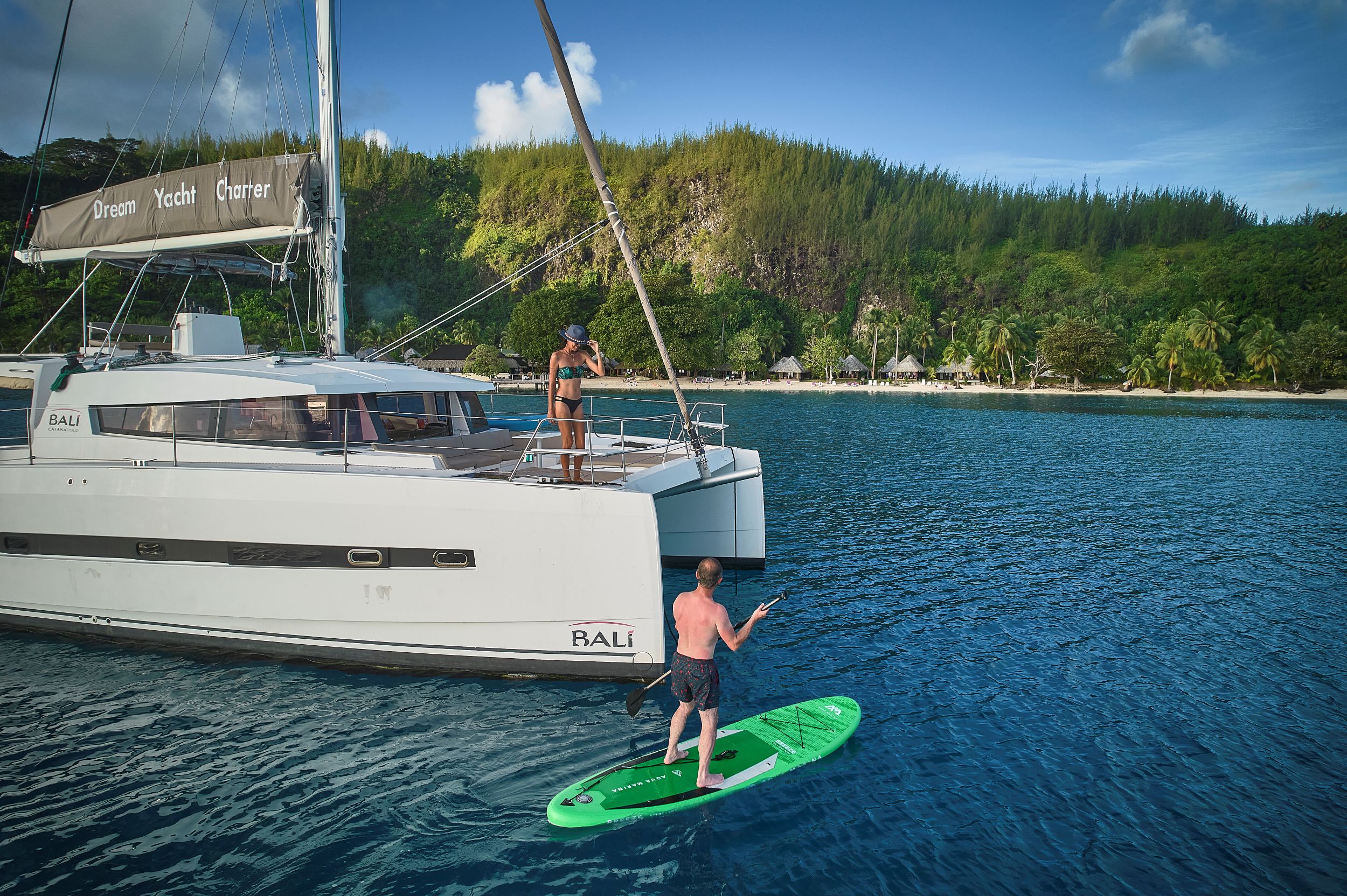 A man on a paddleboard at anchor and a woman standing on the foredeck of a catamaran